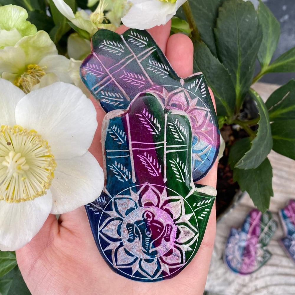 Hand Hamsas mit eingravierten Ganesha Kopf mittig der Handfläche. Dunkles Petrol, dunkles Lila sowie dunkles Grün ziehen sich als drei Streifen über die Hand aus Speckstein.