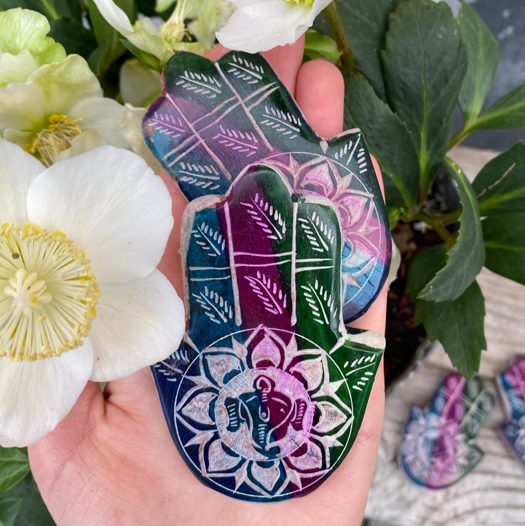 Hand Hamsas mit eingravierten Ganesha Kopf mittig der Handfläche. Dunkles Petrol, dunkles Lila sowie dunkles Grün ziehen sich als drei Streifen über die Hand aus Speckstein.