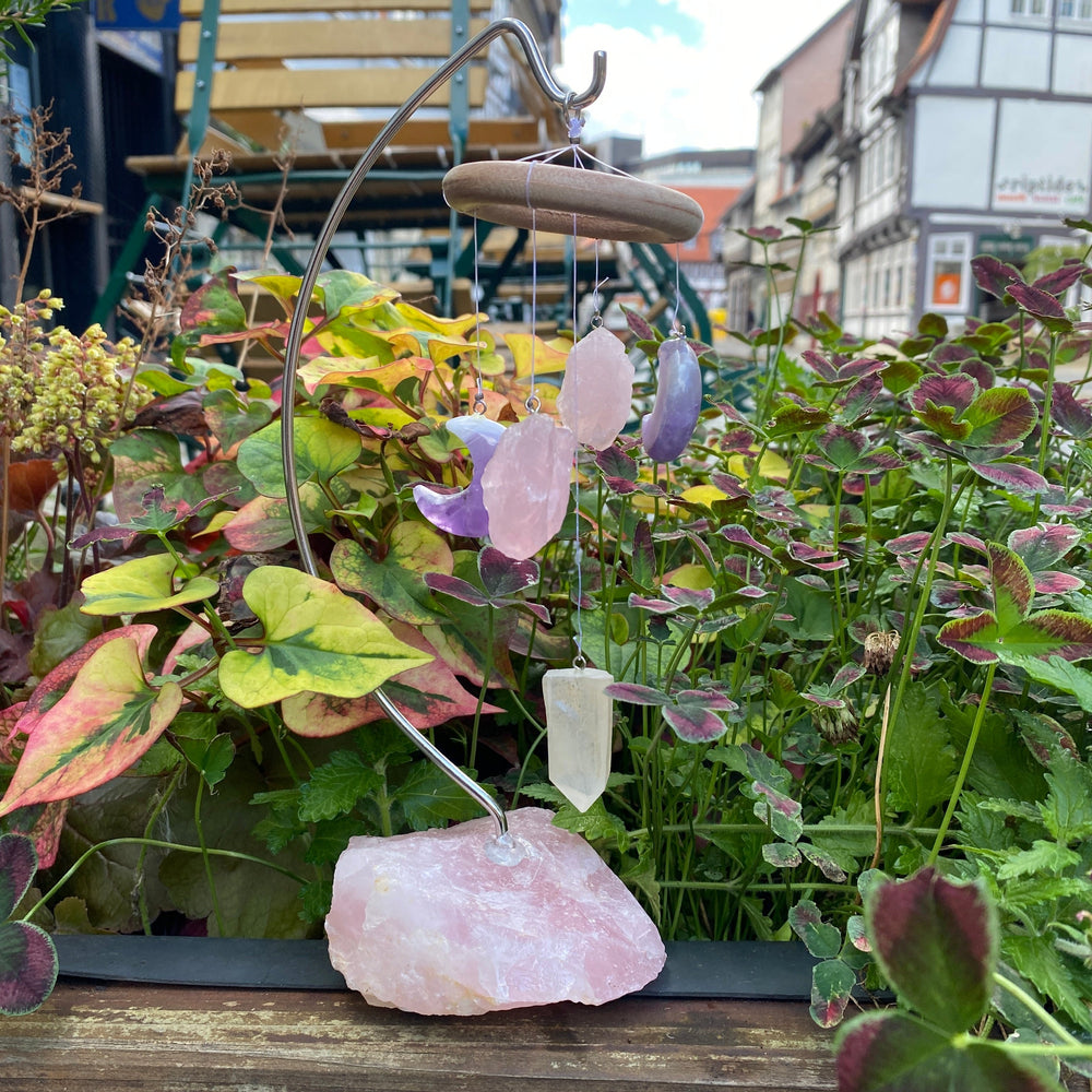 An einem großen Rosenquarzbruchstein ist ein Metallträger, an welchem ein Holzring hängt. An diesem befinden sich zwei Amethyst Monde, zwei Rosenquarz Bruchsteine und ein Bergkristall Pendel. 