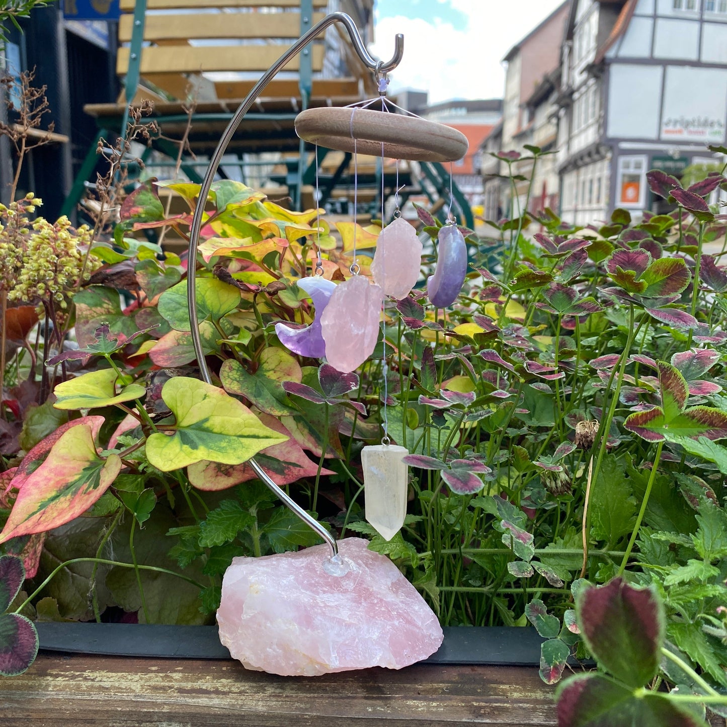 An einem großen Rosenquarzbruchstein ist ein Metallträger, an welchem ein Holzring hängt. An diesem befinden sich zwei Amethyst Monde, zwei Rosenquarz Bruchsteine und ein Bergkristall Pendel. 