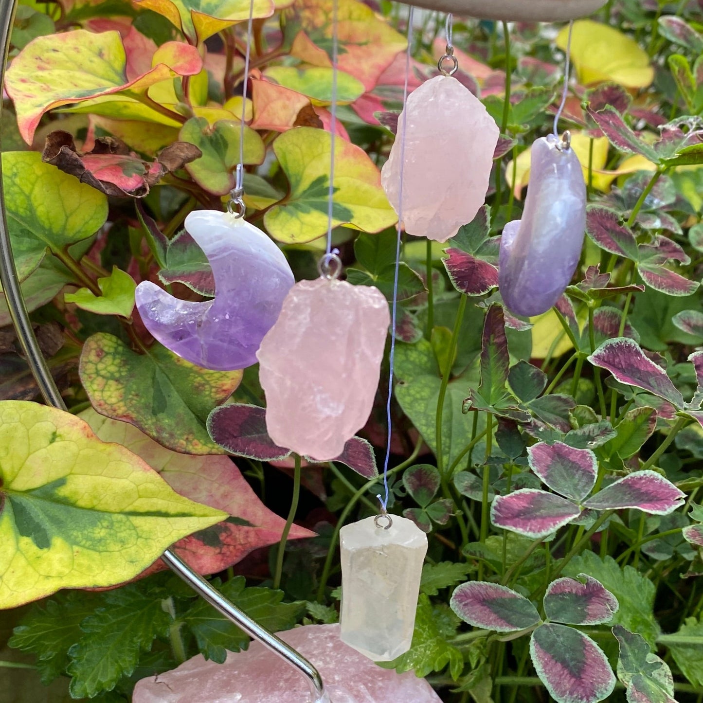 Rosenquarz Bruchsteine, Amethyst Monde und ein Bergkristall Pendel an einem Holzkreis hängend und von einem Rosenquarzfuß gehalten. 
